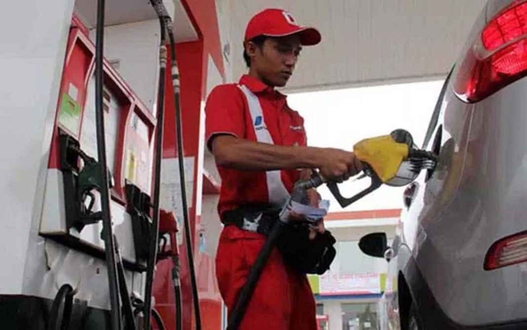 Petugas SPBU sedang melayani pengisian BBM pada kendaraan dengan latar belakang papan harga bahan bakar yang menyala.